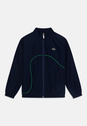 Chaqueta ligera azul marino con cremallera frontal, cuello alto, puños elásticos, líneas curvas verdes como acento y un pequeño logo en el pecho.