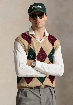 Mężczyzna noszący ciemne okulary przeciwsłoneczne, zieloną czapkę RLX Ralph Lauren, kamizelkę w szkocką kratę, białą koszulę, szare spodnie i zegarek na rękę, stojący z założonymi rękami.