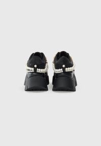 Sneaker nere chunky con catene di perle stratificate avvolte attorno ai talloni, viste dal retro su uno sfondo bianco semplice.