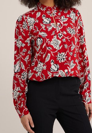 Kobieta ubrana w czerwoną bluzkę z długim rękawem z białymi i czarnymi kwiatowymi wzorami oraz czarne spodnie, przycięta w talii, stojąca na jednolitym tle.