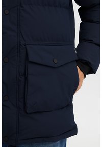 Tmavě modrá péřová bunda s velkou klopovou kapsou zabezpečenou cvočkem. Hladká textura látky a prošité části pro izolaci.