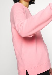 Person, die ein langärmliges pinkes Sweatshirt mit einem seitlichen Schlitz, schwarze Hosen und große Creolen trägt und leicht seitlich schaut.