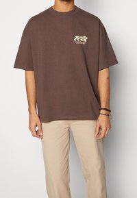 Brązowy, oversize t-shirt wykonany z bawełny, z małym kremowym wzorem na lewej piersi. Krótkie rękawy i okrągły dekolt.