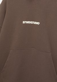 Brązowa bluza z kapturem z przednią kieszenią i białym, pogrubionym napisem "STWDSTUDIO" umieszczonym centralnie nad kieszenią.