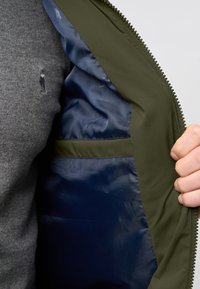 Olivovo zelená bunda s hladkou námorníkovou podšívkou. Interiér obsahuje vrecko a zips. Vonkajšia tkanina vyzerá štruktúrovane a odolne.