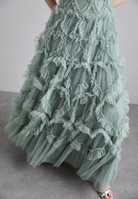 Robe longue verte menthe en tissu transparent avec des accents à volants, des découpes rondes et des motifs à pois à l'ourlet. Sandales argentées visibles.