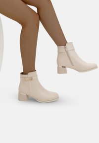 Botines beige hasta el tobillo con un acabado brillante, que presentan un cierre con cremallera, una correa decorativa con un acento dorado y un tacón cuadrado.