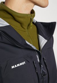 Chaqueta exterior negra con acentos grises y logo de Mammut. Forro de polar verde con cremallera, que presenta un cuello alto y una tela texturizada.