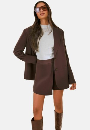 Kvinde iført en brun blazer, matchende asymmetrisk nederdel, hvid top, brune knæhøje støvler og store solbriller mod en ensfarvet baggrund.