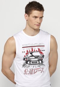 Bijela majica bez rukava s crno-crvenim grafičkim prikazom automobila, uz natpis "TOKYO" i japanski tekst. Mekan pamučni materijal i ležeran kroj.