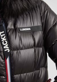 Czarna kurtka puchowa z błyszczącym, pikowanym wykończeniem. Posiada sztuczny kołnierz z futra, paski na zamku błyskawicznym oraz prostokątną naszywkę z napisem "JACKIT."