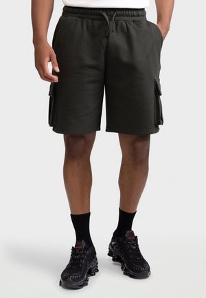 Personne portant un short cargo noir avec des poches latérales, des chaussettes noires et des chaussures de sport noires, debout devant un fond clair uni.