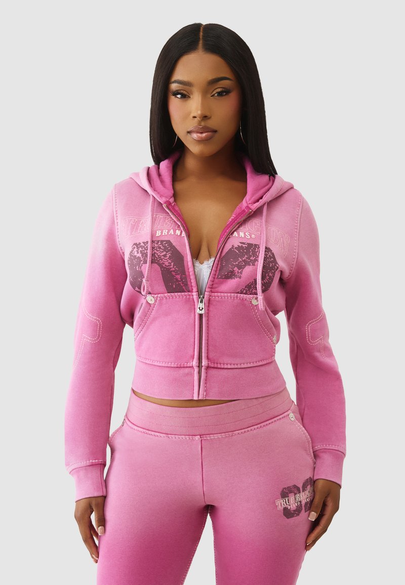 Hoodie rose zippé avec poches avant et design graphique, associé à un pantalon de survêtement assorti. Effet ombré passant du clair au foncé. Texture de tissu douce.