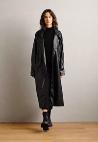 Černý trenčkot vyrobený z lesklého materiálu, s dvouvrstvým designem, velkým límcem a dlouhými rukávy, nošený přes černý outfit.