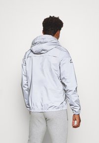 Light grey, hooded jacket with smooth, reflective fabric. Features elastic cuffs and a cinched waist, with a back yoke and logo near shoulder.