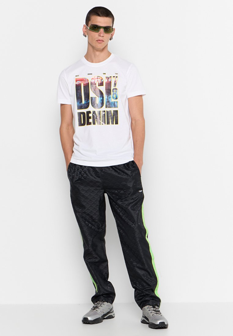 T-shirt bianco con grafica colorata "DSL DENIM", abbinato a pantaloni da pista neri a motivo romboidale con strisce verdi ai lati e sneakers grigie.