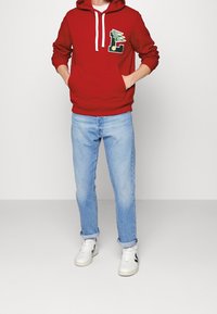 Personne portant un sweat à capuche rouge avec poche avant et écusson, un jean bleu clair retroussé, et des baskets blanches avec des détails noirs, debout devant un fond uni.