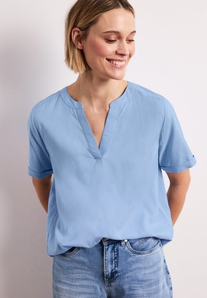 Femme aux cheveux blonds courts souriante, regardant vers le bas, portant un chemisier à manches courtes bleu clair et un jean bleu sur un fond uni.