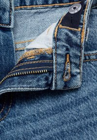 Denimová látka v modré barvě s knoflíkovým zapínáním, odhaleným zipem se zlatými zuby a kontrastním žlutým šitím, s třepenými okraji podšívky kapes.