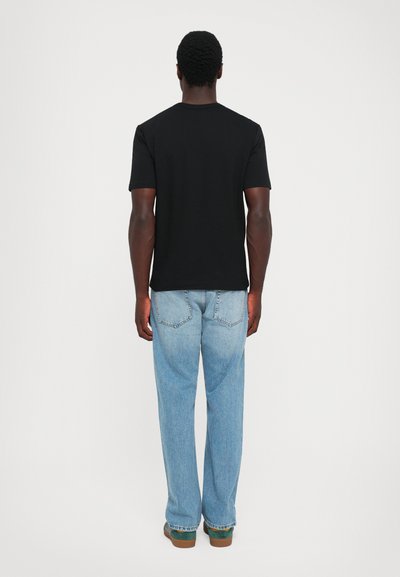Man standing facing away wearing a black t-shirt, light blue jeans, and green shoes on a plain white background.