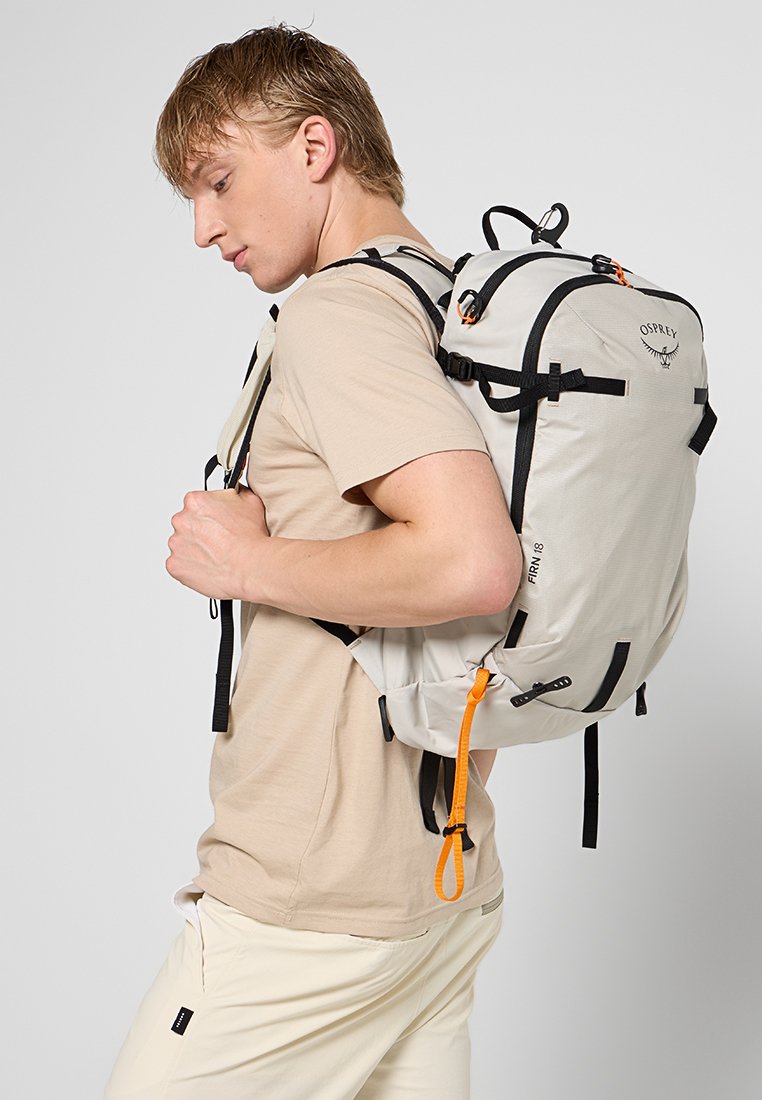 Mochila gris claro con detalles en negro y cremalleras naranjas, con un diseño redondeado, correas ajustables y múltiples puntos de sujeción.