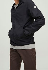 Dark blue hooded jacket with a zip closure, side pockets, and a light gray hem, featuring a circular logo on the left sleeve.