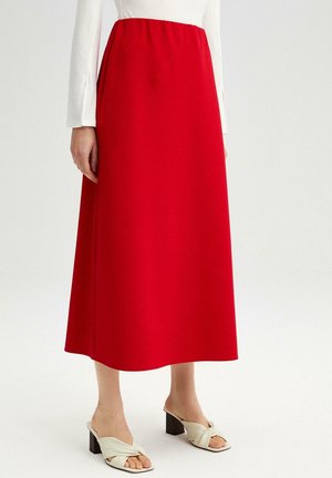 Kvinde iført en lang, rødhøj taljeskørt kombineret med hvide hælede sandaler og en hvid bluse med lange ærmer, stående mod en ensfarvet baggrund.