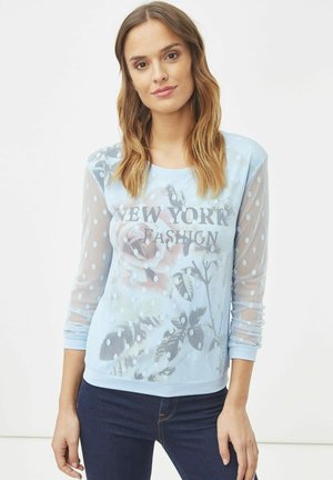 Top de manga larga azul claro con un diseño transparente de lunares y estampado floral, con el texto "FASHION DE NUEVA YORK" en el centro.