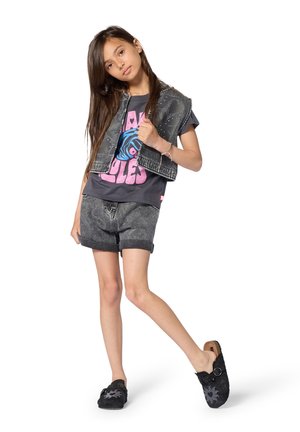 Gilet in denim grigio indossato sopra una t-shirt grigio scuro con grafiche rosa, abbinato a pantaloncini grigi e scarpe slip-on nere con un motivo di sole.