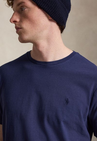 Νέος άνδρας που φοράει σκούφο από πλεκτό ύφασμα σε ναυτικό μπλε και αντίστοιχο t-shirt με στρογγυλή λαιμόκοψη σε ναυτικό μπλε, με μικρό κεντημένο λογότυπο στο στήθος.