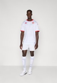Balta futbola krekla ar sarkanām akcentiem, ar rakstainu dizainu, atbilstošas baltas šortus, ceļa garuma baltas zeķes un melni futbola apavi.