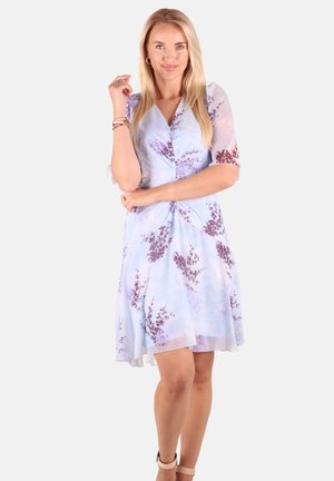 Frau mit langen blonden Haaren, die ein hellblaues geblümtes Kleid und beige High Heels trägt, steht lächelnd vor einem einfarbigen Hintergrund.