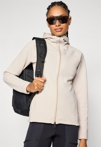 Veste beige à capuche avec une finition texturée, fermeture éclair et détail de logo. Portée avec un sac à dos noir et des lunettes de soleil.