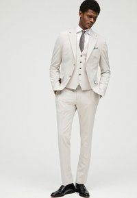 Traje de tres piezas en beige claro, con chaqueta y chaleco a medida, combinado con una camisa blanca y una corbata estampada, completado con zapatos negros.