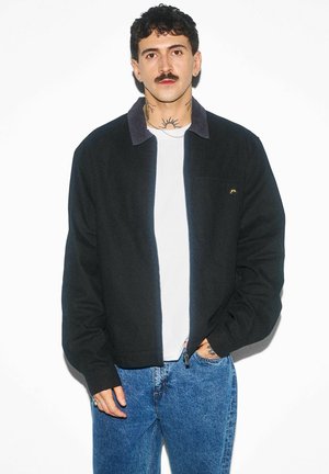 Mężczyzna z kręconymi włosami i wąsami, ubrany w czarną kurtkę na białej koszuli i niebieskich dżinsach, stojący na białym tle.