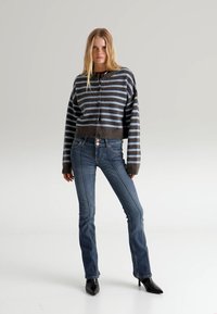 Obrezan pulover z gumbi, temno rjave in modre vodoravne črte, kombiniran z oprijetimi modrimi kavbojkami in črnimi škornji z visoko peto.