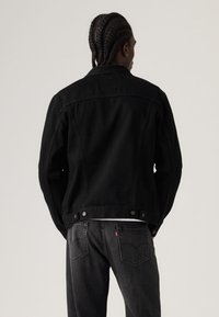 Čierna rifľová bunda so štruktúrovaným dizajnom, ktorá má zadný diel, patentové gombíky a hladkú textúru. Nosí sa so sivými džínsami.