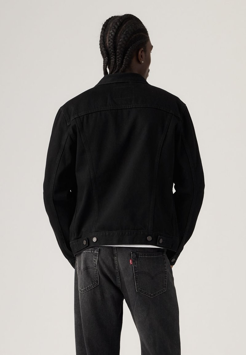 Black denim jacket with a structured design, featuring a back yoke, snap buttons, and a smooth texture. Worn with grey jeans.