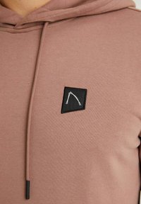 Mikina s kapucí vyrobená z měkké, světle růžové látky, se černým čtvercovým štítkem s bílým geometrickým vzorem na hrudi. Kordu jsou černé.