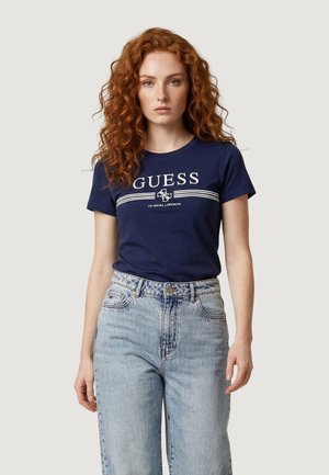 Femme aux cheveux roux portant un t-shirt GUESS bleu marine et un jean taille haute bleu clair, debout devant un fond gris clair uni.