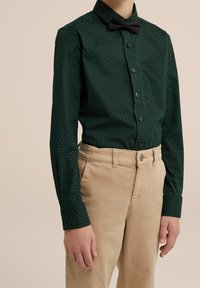 Grön prickig skjorta med knäppning och svart fluga, i kombination med ljusbeiga byxor. Skjortan har en bröstficka och långa ärmar.