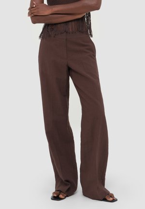 Person iført højtaljede, løse brune bukser, sorte sandaler med firkantet tå og en mørk crop top med frynser, stående med armene krydset.