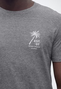 Κοντινή λήψη γκρι ριγέ μπλουζάκι με λευκό λογότυπο φοίνικα και κείμενο "EST. 07 Denim Design Studio" στο στήθος.