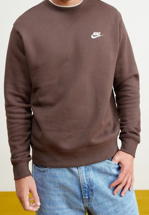 Man draagt een bruine Nike-trui gecombineerd met lichtblauwe spijkerbroeken, met één hand in de zak en een effen achtergrond.