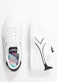 Bílé tenisky s černými akcenty, hladký syntetický svršek, perforovaný design, texturovaný černý podpatek a barevná vzorovaná stélka.