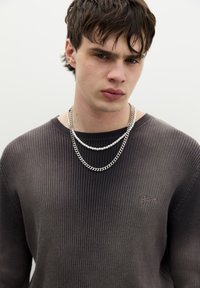 Grijze geribbelde trui met een ronde hals, gelaagd met twee kettingen: een zilveren schakelketting en een witte kralenketting.