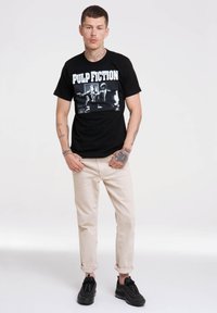 Sort bomulds t-shirt med "PULP FICTION" tekst og et grafisk print. Bæres med beige optrullede bukser og sorte sneakers.