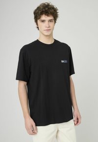 T-shirt en coton noir à manches courtes, col rond, avec un petit logo sur le côté gauche de la poitrine. Texture lisse, coupe décontractée.