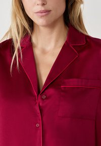 Bordowy satynowy top piżamy z zapięciem na guziki, kołnierzem typu notch i jedną kieszenią, z kontrastowym obszyciem wzdłuż krawędzi.