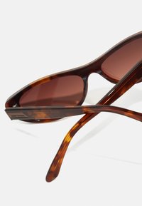 Gafas de sol de carey con lentes marrones, que presentan un diseño elegante y angular y un marco de plástico suave. Incluyen patillas delgadas con detalles texturizados.